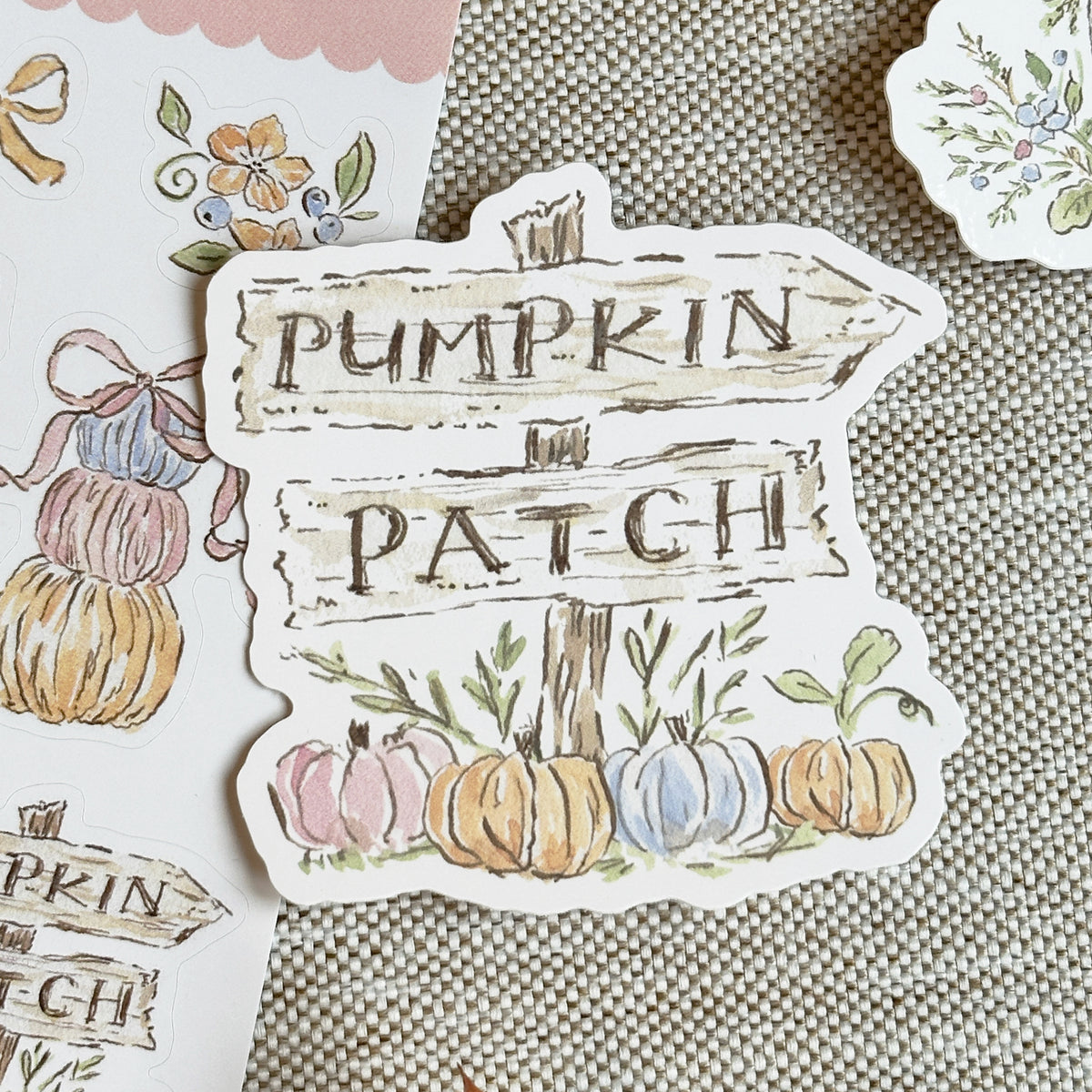 A die-cut sticker featuring a rustic wooden signpost that reads 'PUMPKIN PATCH' with watercolor illustrations of pastel pumpkins and green leaves, shown on a textured beige fabric background.