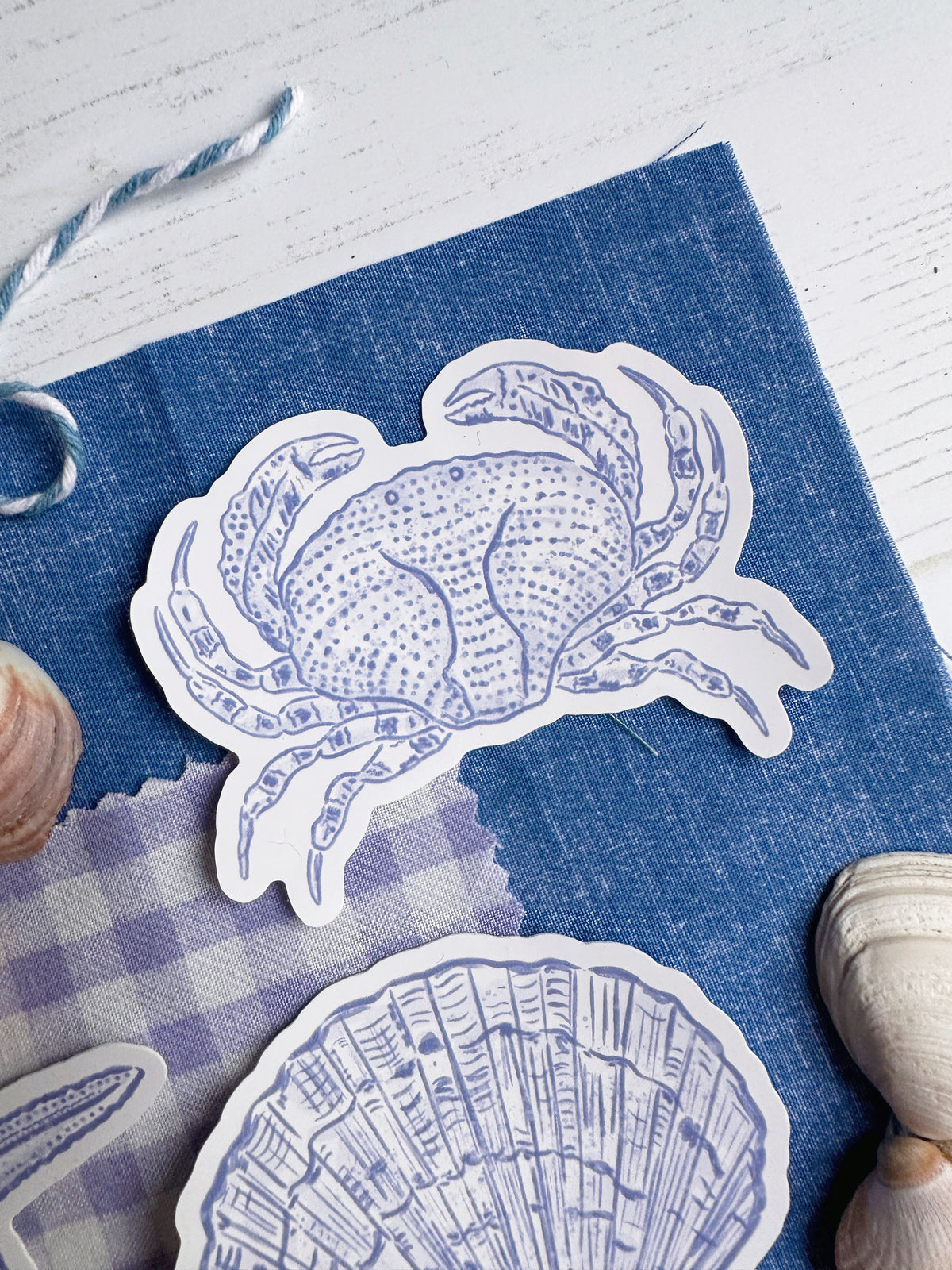 A close-up of a blue fabric surface with die-cut paper stickers featuring a blue crab and a blue seashell outline, styled with small seashells and a gingham cloth.