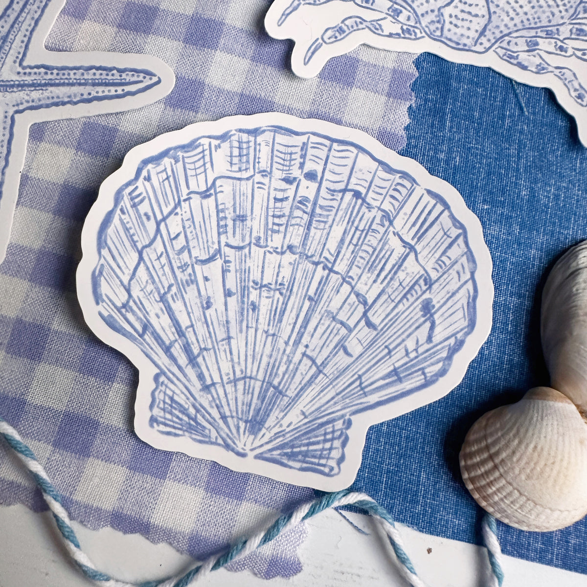 A die-cut sticker shaped like a scallop shell, printed with a blue and white illustration of open books, displayed on a blue and white gingham fabric alongside other seashell-shaped stickers and small real seashells.