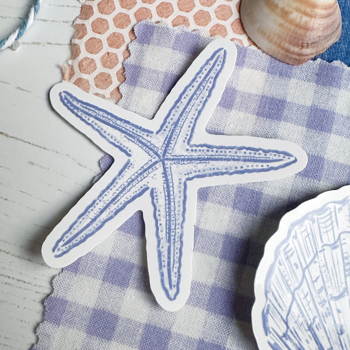 A die-cut starfish-shaped sticker or decal with a blue line drawing on a white background, placed on a purple-and-white gingham cloth with nautical props around it.