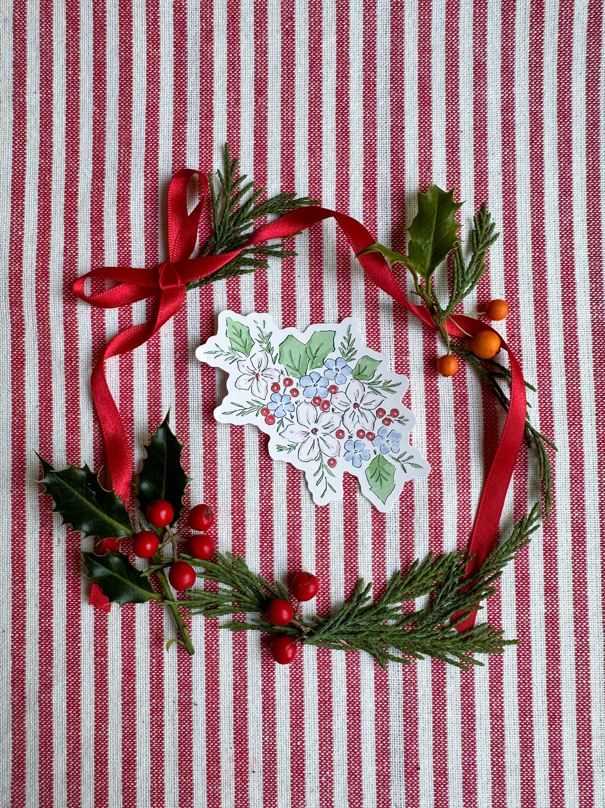 A Christmas-themed flat lay featuring a green pine and holly garland with red berries, a red ribbon bow, and a small white paper ornament printed with a floral design, all arranged on a red and white striped fabric background.