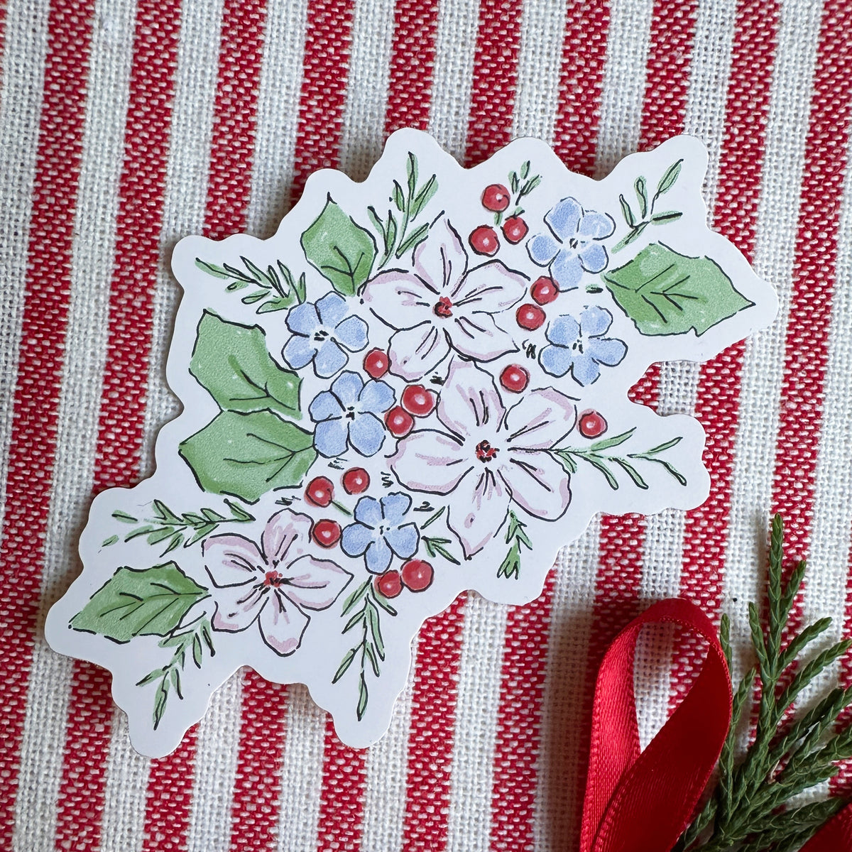 A die-cut decorative ornament or sticker featuring a hand-painted floral bouquet with blue, pink, and white flowers, green leaves, and small red berries, placed on a red and white striped fabric background with a red ribbon and evergreen sprig.