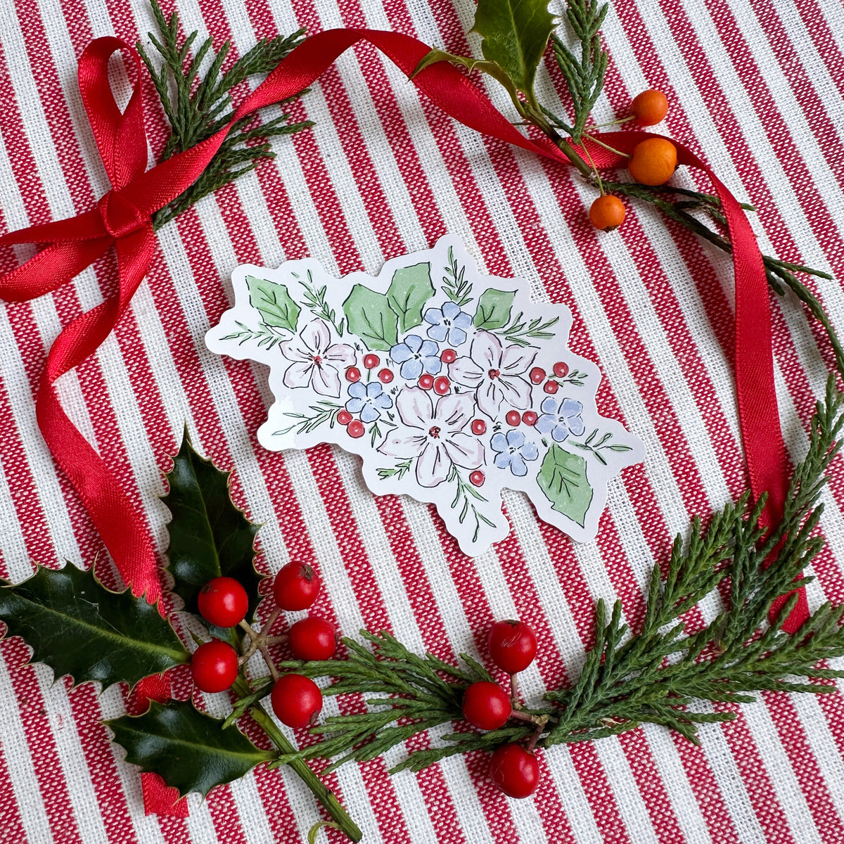 A die-cut sticker featuring a winter floral illustration with white flowers, blue blossoms, green leaves, and red berries, arranged on a red and white striped fabric with holly leaves, pine branches, and red ribbon.