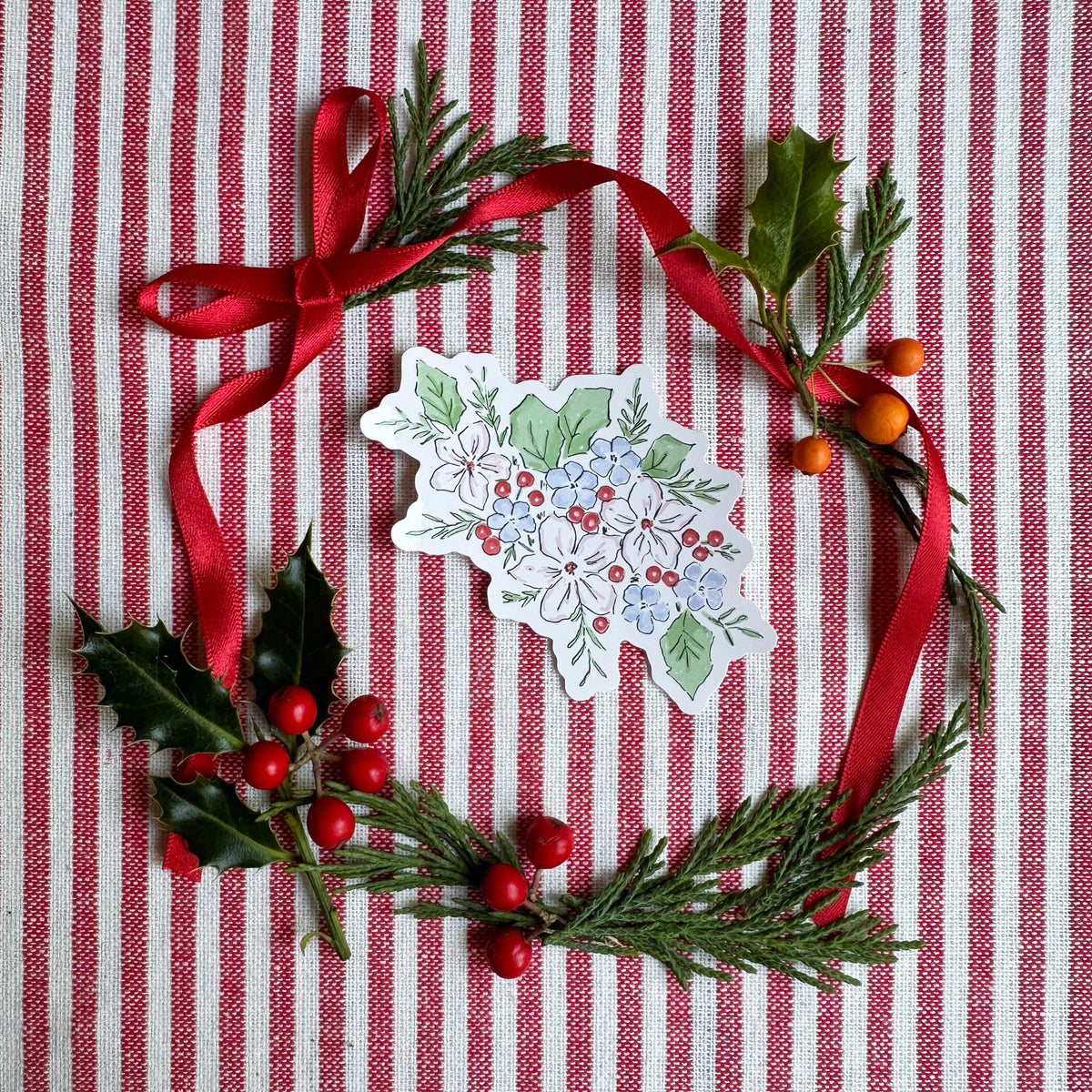 A hand-painted Christmas ornament shaped like a holly leaf, decorated with blue and white flowers, red berries, and green leaves, arranged in a wreath with a red ribbon on a red and white striped fabric background.
