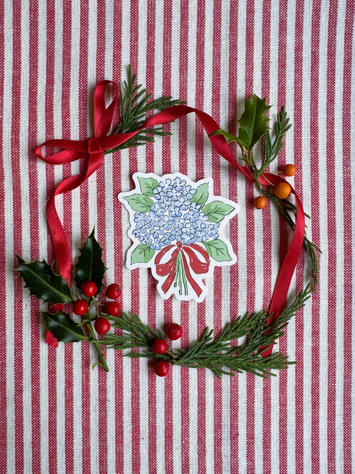 Vinyl die cut sticker of illustrated hydrangea bouquet with red bow. The background is red and white striped fabric with red ribbon greenery and berries.