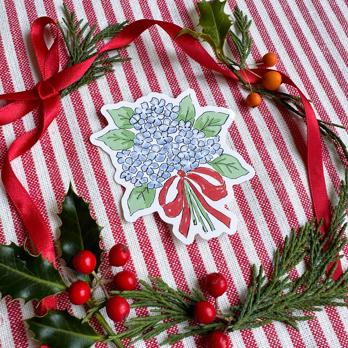 Vinyl die cut sticker of illustrated hydrangea bouquet with red bow. The background is red and white striped fabric with red ribbon greenery and berries.