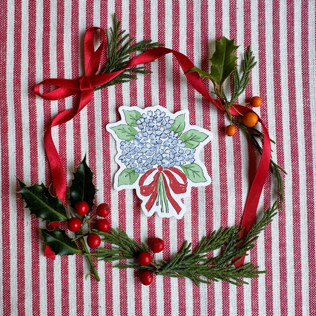 Vinyl die cut sticker of illustrated hydrangea bouquet with red bow. The background is red and white striped fabric with red ribbon greenery and berries.