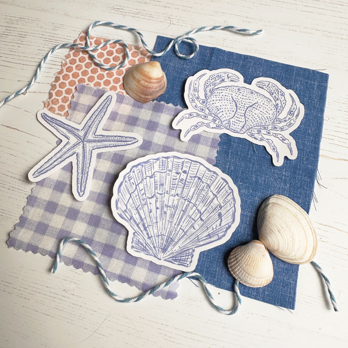 A styled flat-lay of nautical-themed paper stickers, including a starfish, scallop shell, crab, and clam, arranged on blue and lavender gingham fabric with seashells and twine on a white wood background.