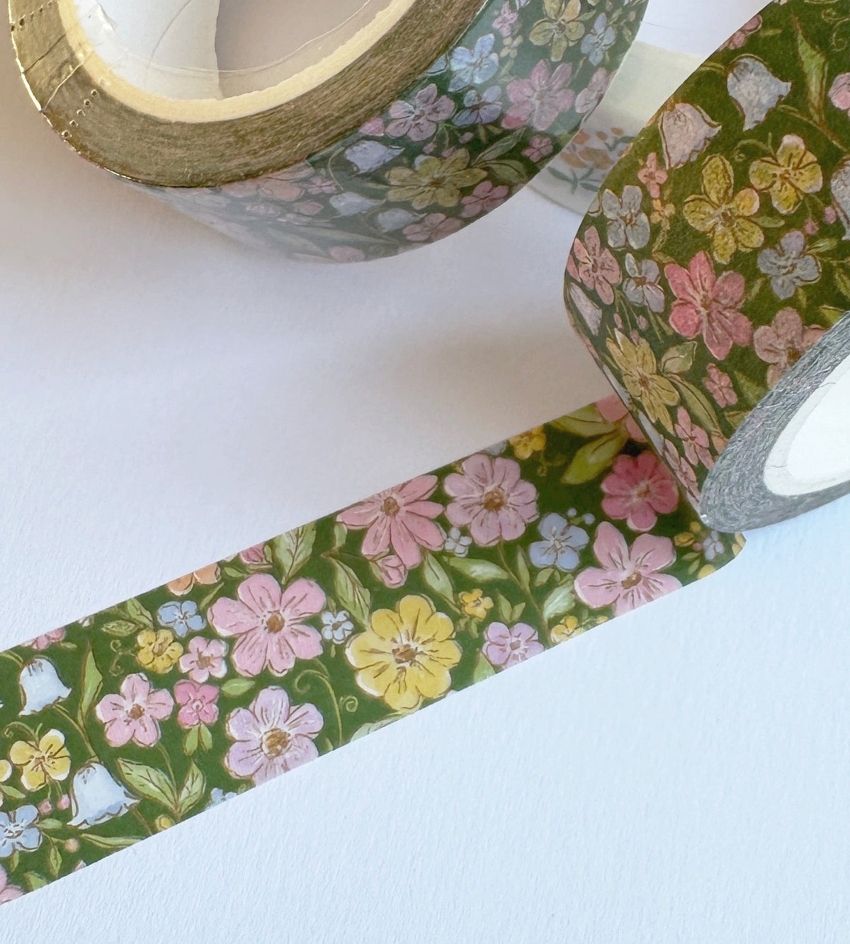 Three rolls of floral washi tape on a white background, with one roll unrolled to show the pattern. The tape features a green background with pink, yellow, and light blue flowers.