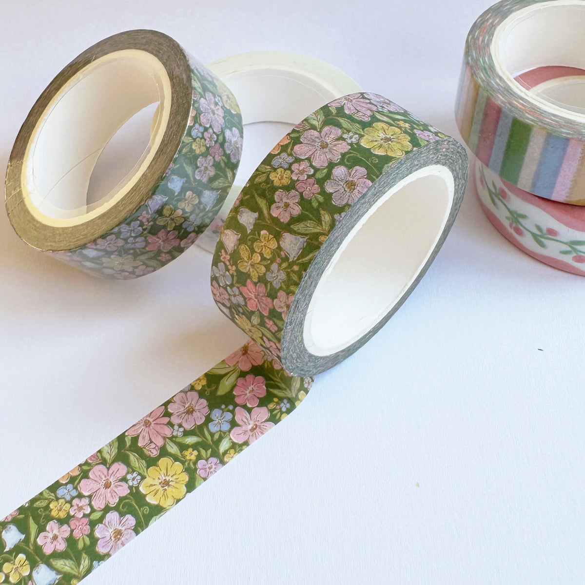 A set of washi tapes with floral and striped patterns, displayed on a clean white background. One roll is partially unrolled to show the pattern, which features pink, yellow, and green flowers on a dark green background.