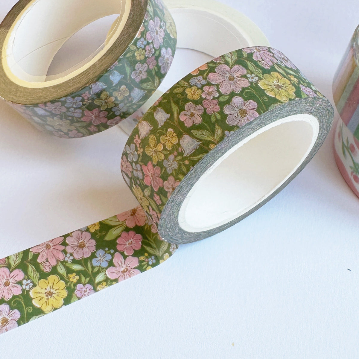 Floral washi tape rolls on a white background, featuring a green base with pink, yellow, and blue flowers. One roll is unrolled to show the pattern and gray backing.